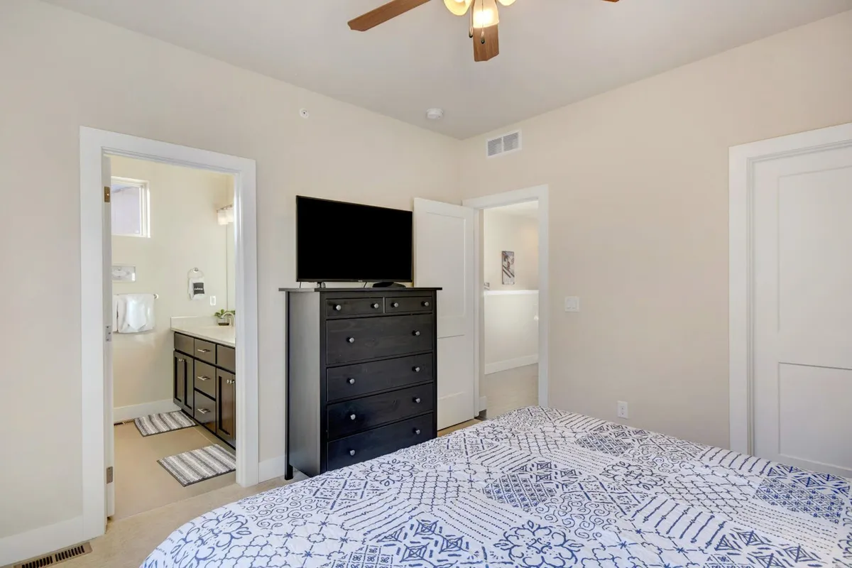 A spacious bedroom with a large bed featuring patterned bedding, a dresser with a television, and natural light pouring in through the windows. Doorways lead to a bathroom and a hallway, creating a sense of privacy and comfort ideal for restful nights and easy mornings.