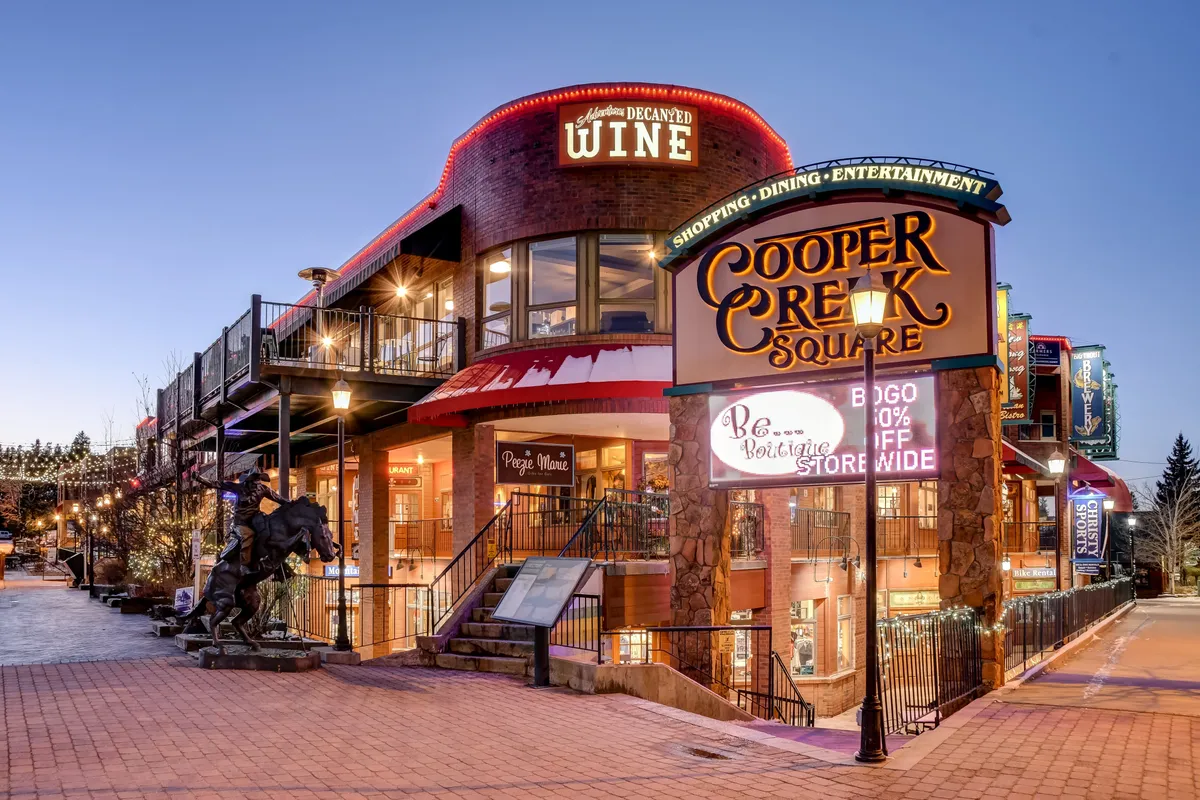 A bustling exterior scene at Cooper Creek Square, with its distinctive sign, inviting wine shop, festive lights, and people enjoying shopping and dining. The setting feels lively and welcoming, capturing the vibrancy of downtown Winter Park.