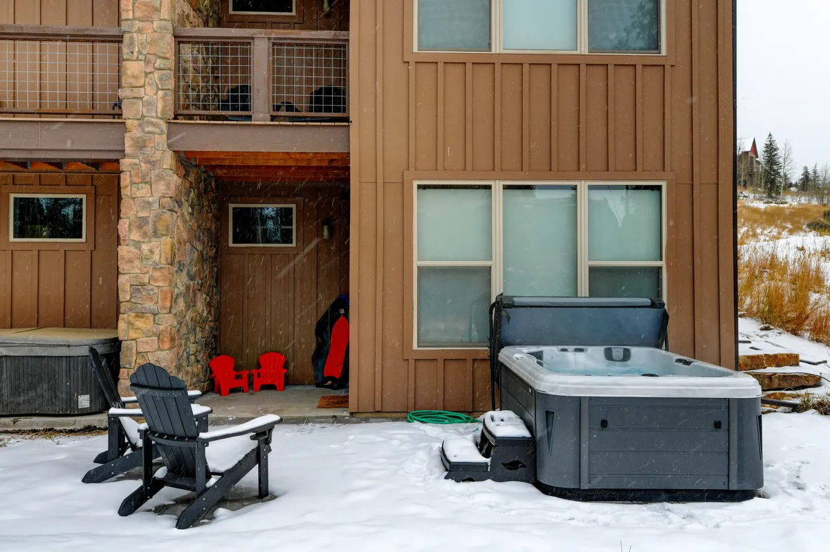 A cozy exterior setting shows a steaming hot tub surrounded by Adirondack chairs, set against a backdrop of a stone-clad building and a snow-blanketed ground. The scene exudes warmth and relaxation, inviting guests to unwind after a day of outdoor exploits.