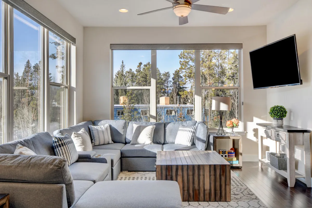 The image showcases a spacious, sunlit living room with a plush sectional sofa, a stylish wooden coffee table, and large windows offering serene wooded views. The inviting setup is perfect for relaxing, gathering with family and friends, or enjoying a cozy movie night in the mountains.