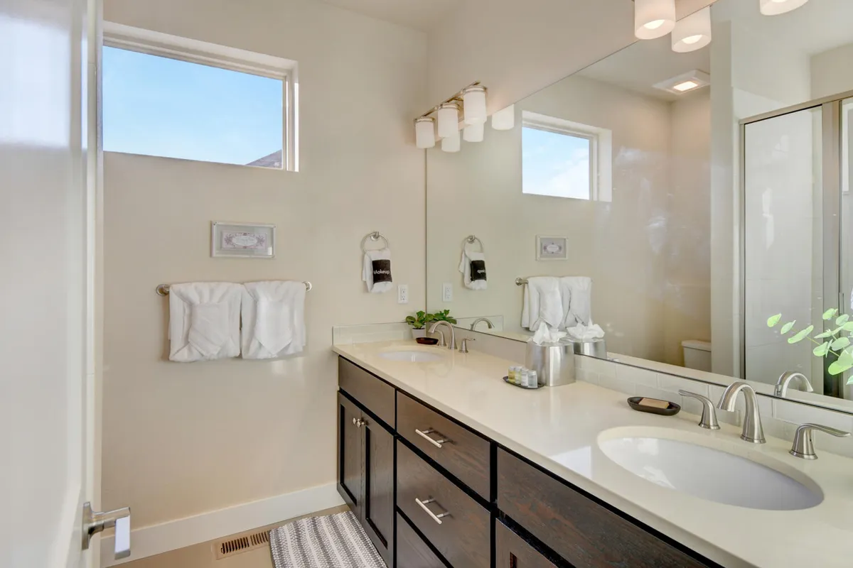 A calming bathroom with double vanities, a large mirror, and abundant natural light. The simple, clean decor and visible spa-quality linens evoke a sense of serene self-care and luxury.
