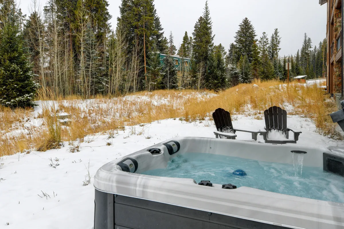 An outdoor hot tub surrounded by snow-covered ground and tall trees, with wooden chairs nearby. The setting is tranquil, with the hot tub steaming against a backdrop of wintry nature, creating a sense of privacy and relaxation.