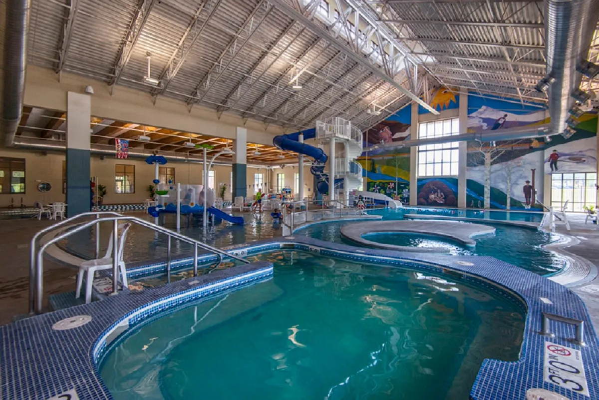 A lively indoor pool area with colorful water slides, children laughing and playing in the water, and vibrant murals decorating the walls. The scene feels energetic and joyful, capturing families enjoying the rec center's aquatic amenities.